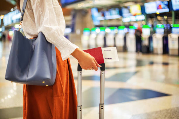 Woman holding passport and boarding pass, standing beside suitcase in airport terminal, reflecting travel and passport services.