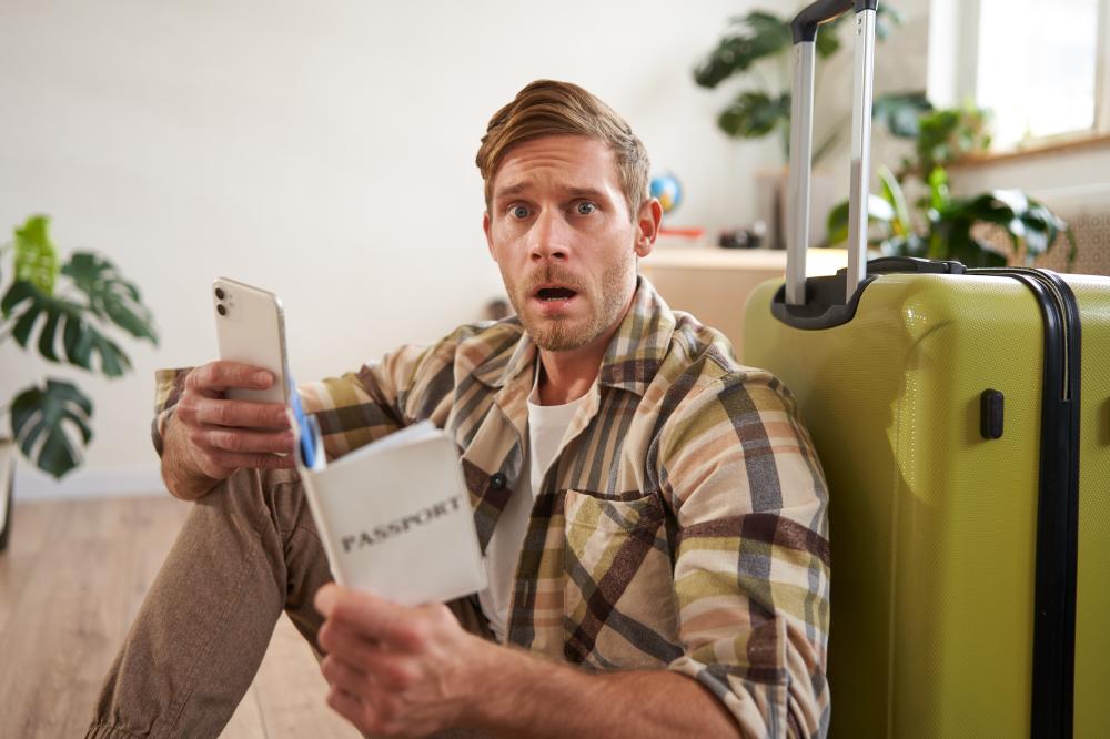 Man with a shocked expression sitting on the floor next to a suitcase, holding a passport and smartphone, reflecting urgency related to emergency passport services in Phoenix.